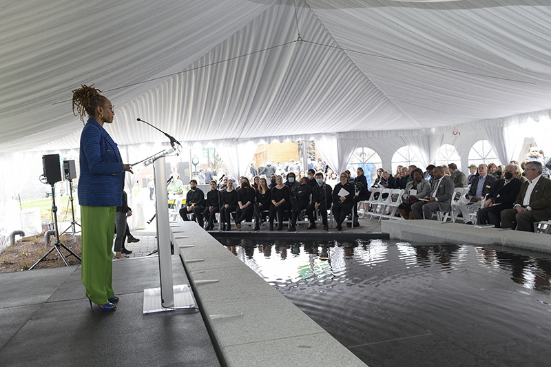 Wendi Manuel Scott in a blue blazer and green pants stands at a lectern in front of the fountain at the Enslaved People of George Mason Memorial, addressing the seated crowd at the dedication ceremony
