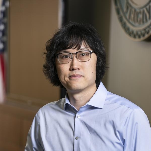 A photo of CS associate professor Lap-Fai (Craig) Yu, wearing a light blue button-up shirt, sitting indoors with an American flag blurred in the background.