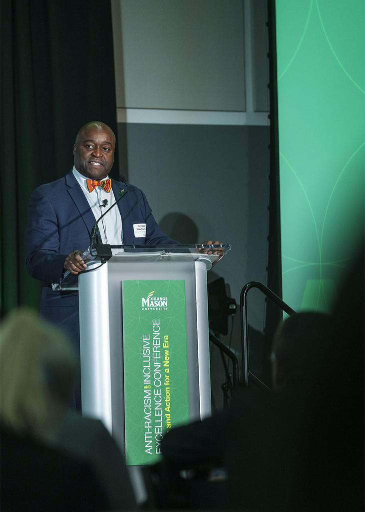 Mason president Gregory Washington stands at a lectern, a green sign reads Anti-Racism Inclusive Excellence Conference. Dr. Washington is wearing a red bowtie and a dark blue suit.