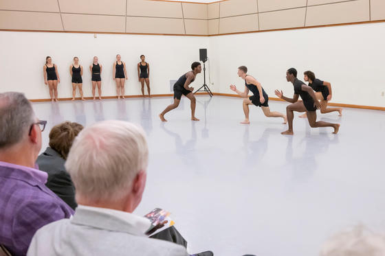 Dancers wearing back perform in a light-filled dance studio in front of an audience.