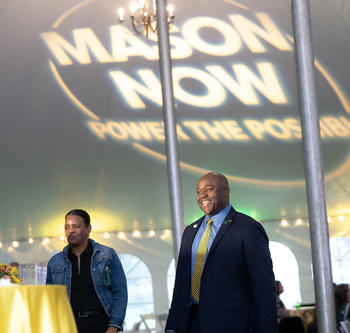 President Washington stands inside a white event tent. The Mason Now logo is projected onto the tent ceiling behind him.